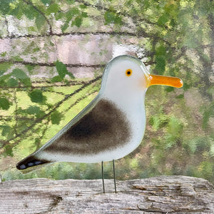 A decorative seagull glass figurine with a white head and body, a dark brown back, and a bright orange beak, standing on thin metal legs against a blurred outdoor background.