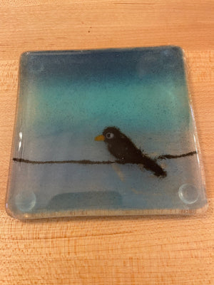 A square glass coaster featuring a scene of a small black bird perched on a branch against a blue and teal gradient background, shown on a light wood surface.