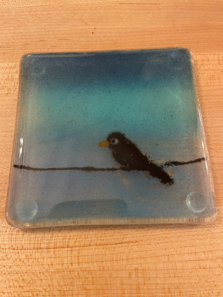 Charger l'image dans la galerie, A square glass coaster featuring a scene of a small black bird perched on a branch against a blue and teal gradient background, shown on a light wood surface.
