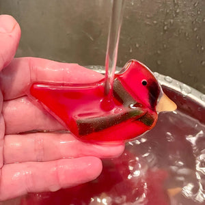 A red glass bird wearing a green glass scarf is being held in the hand and washed under a stream of water.