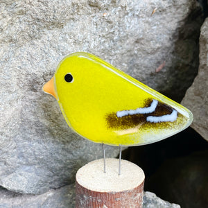 Yellow glass bird ornament on a wooden base against a stone background