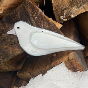 A white bird-shaped hanging ornament with a glossy finish and small black eye, suspended against a background of stacked firewood and a bit of snow.