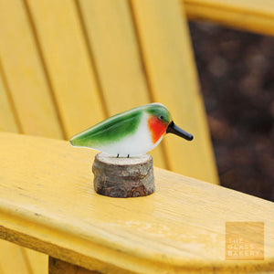 a red and green ruby throated hummingbird sits on a log. the log is on a bright gold painted Adirondack chair