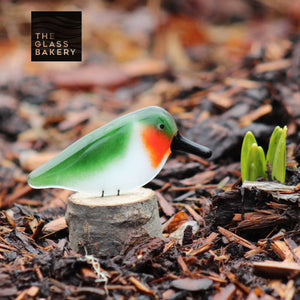 A tiny glass ruby throated hummingbird sits on a small log in front of some daffodils which are just emerging from the ground.