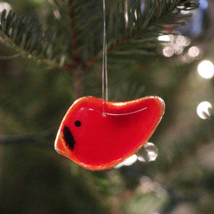 A small red glass bird in the shape of a Cardinal, hangs from a Christmas Tree. You can see the Christmas lights in the background.