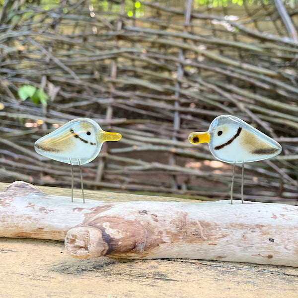 Load image into Gallery viewer, a pair of brown and white fused glass piping plovers are perched on driftwood, in front of a background of branches
