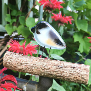 a fused glass mini chickadee bird sits on a small log in amongst bee balm