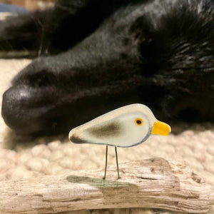 a small glass mini seagull perches on driftwood in front of a black dog. They are both on a wool rug