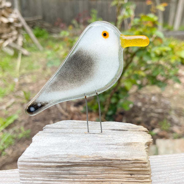 Load image into Gallery viewer, Glass seagull bird sculpture with a yellow beak on a wooden driftwood perch outdoors
