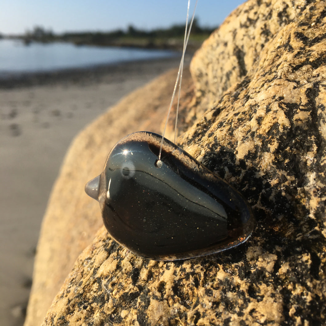 A tiny black shiny glass crow is hanging in front of a rock on a beach.