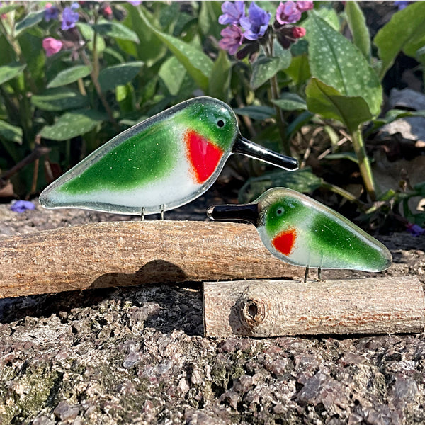 Load image into Gallery viewer, Two green and red hummingbird glass birds on a wooden log with a natural background of plants and flowers.
