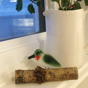A green and red glass hummingbird sits on a windowsill next to a white jug full of holly with red berries