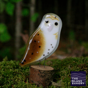 A shiny brown speckled glass owl sits on a log which is standing on moss. Behind is a dark forest.