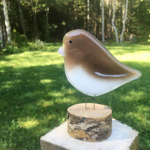 Brown and white glass bird ornament with pink beak. The bird sits on a slice of log for a perch and the backdrop is a green wooded landscape