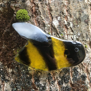 Close up image of a glossy black and gold glass bee with white wing. The bee is hanging in front of a tree.