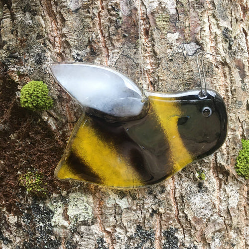 Close up image of a glossy black and gold glass bee with white wing. The bee is hanging in front of a tree.