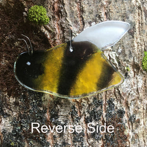 Reverse of close up image of a glossy black and gold glass bee with white wing. The bee is hanging in front of a tree.