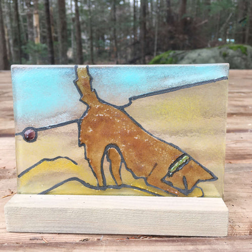 Fused glass panel featuring a brown dog in a green collar, digging for a purple ball in the sand on a beach.