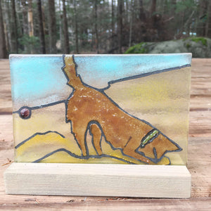 Fused glass panel featuring a brown dog in a green collar, digging for a purple ball in the sand on a beach.