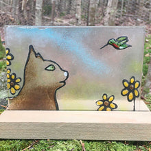 Load image into Gallery viewer, Glass picture tile featuring a brown cat with green eyes, watching a ruby throated hummingbird. the cat is sitting amongst yellow and brown daisys
