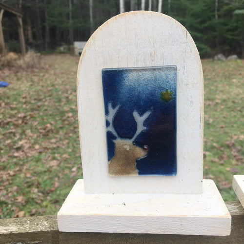 An ivory coloured wooden arch-shaped stand with a fused glass tile on the front. The tile features a reindeer looking at a star in a midnight blue sky.