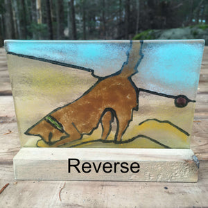 Reverse side of a fused glass panel featuring a brown dog in a green collar, digging for a purple ball in the sand on a beach.