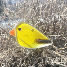 Load image into Gallery viewer, a yellow and black glass female goldfinch hanging ornament dangles in front of a frosty bush

