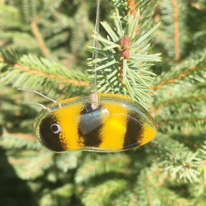 A small gold and black fused glass bee hangs in front of an evergreen tree