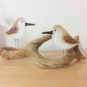 a pair of brown and white glass sandpiper birds are perched on a piece of wriggly driftwood. The driftwood is placed on a wooden table.