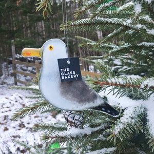 A grey, white and black glass seagull hanging bird hangs from a snowy Christmas tree. 