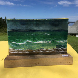 A glass picture tile in a pine slotted stand. The glass depicts a coastal scene with choppy seas, wet sand and moody skies.