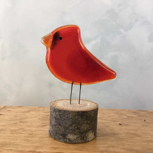 A glossy small red glass cardinal bird ornament is perched on a log which stands on a wooden table.
