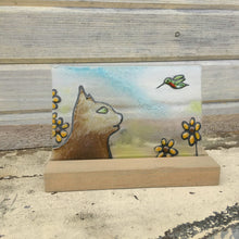 Load image into Gallery viewer, Art glass tile featuring profile of a brown cat with green eyes watching a hummingbird in amongst the yellow brown eyed susan flowers. Glass tile.
