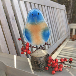 A blue and tan glass bluebird ornament is perched on a grey-washed garden bench with rowan berries around his feet.