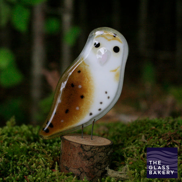 Charger l&#39;image dans la galerie, A shiny brown speckled glass owl sits on a log which is standing on moss. Behind is a dark forest.
