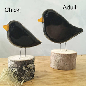 Two fused glass blackbirds with yellow beaks sit on logs. They are on a table.