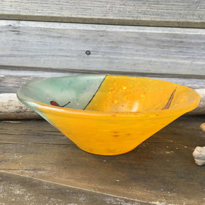 The side view of a conical shaped fused glass dish which is primarily orange and aqua. The dish sits on a wooden mantle