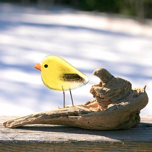 A small glass yellow and black goldfinch bird ornament perches on a piece of driftwood in front of a snowy background