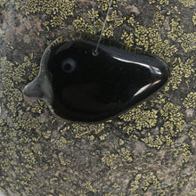 Load image into Gallery viewer, A small glossy black glass crow with a grey beak. Hanging ornament placed on a lichen covered  rock
