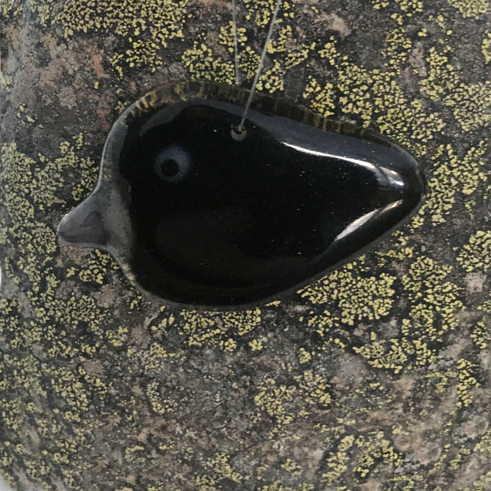 A small glossy black glass crow with a grey beak. Hanging ornament placed on a lichen covered  rock
