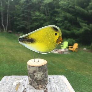 a small yellow and black goldfinch bird ornament perches on a log in front of a grassy lawn background