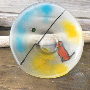 A round fused glass dish, half blue, half yellow featuring a brown dog with a ball playing on a beach