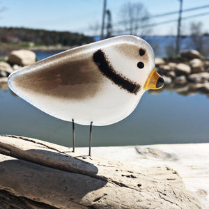 A fused glass plover ornament sits on a driftwood perch. Behind the bird is an out of focus photo of a pond.