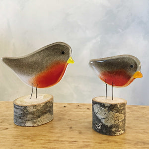 In the foreground are two different sized brown and red glass robin bird ornaments perched on logs on a wood table. The background is a painted wall.