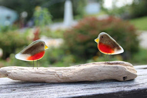 In the foreground are two brown and red glass robin bird ornaments perched on a driftwood log. The background is out of focus but a garden scene.