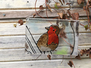 A glass tile featuring a British Robin. The tile hangs against a natural backdrop of winter clematis. 