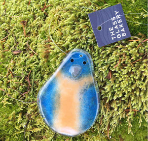 blue and flesh toned fused glass bird faces the camera. he's laying on some moss.