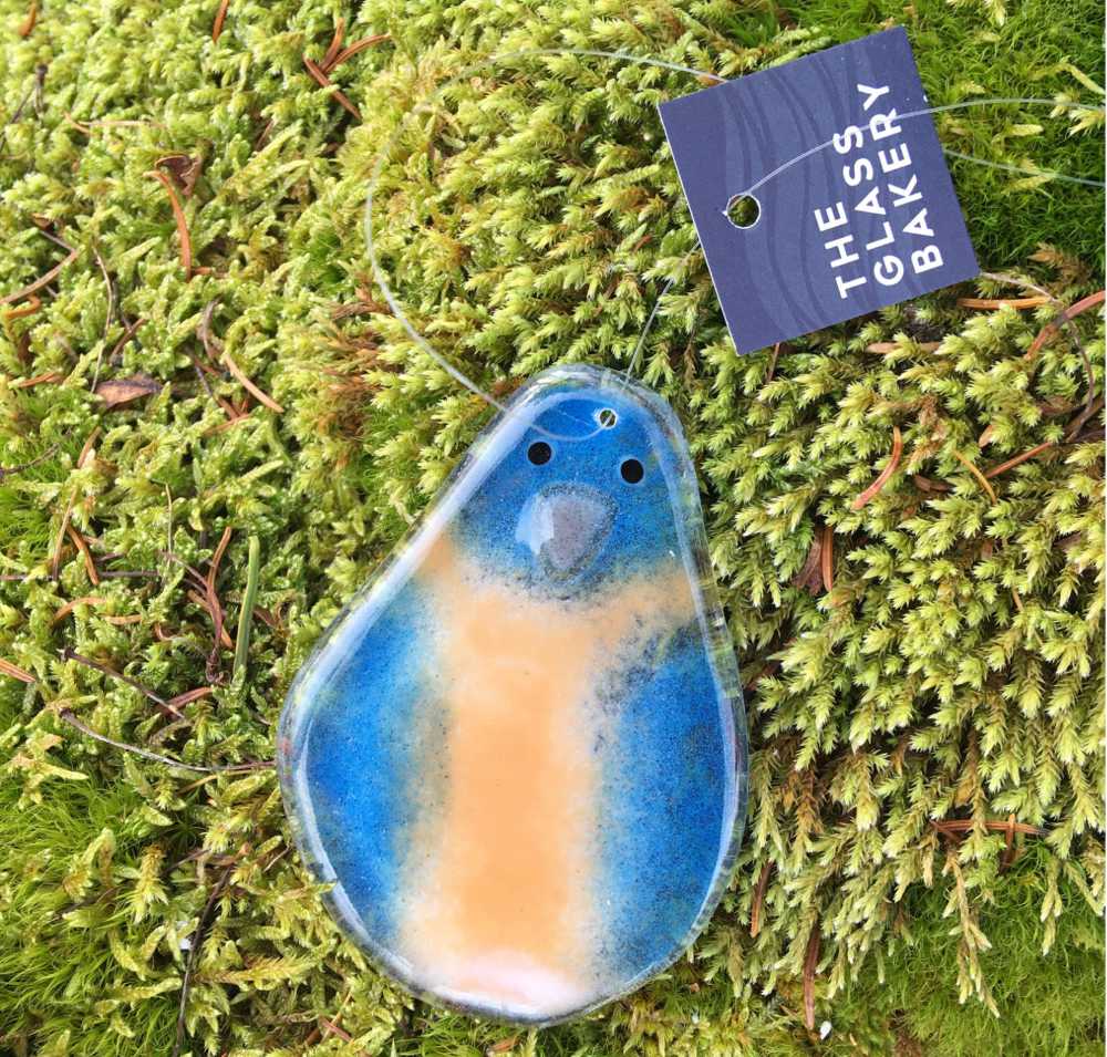 blue and flesh toned fused glass bird faces the camera. he's laying on some moss.