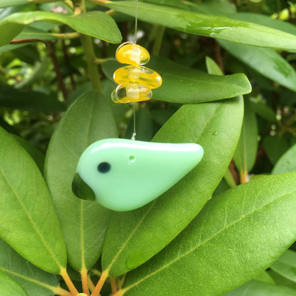 Load image into Gallery viewer, a two toned fused glass green Bird hanging bird is accented by yellow glass beads. The bird is on rhododendron leaves.
