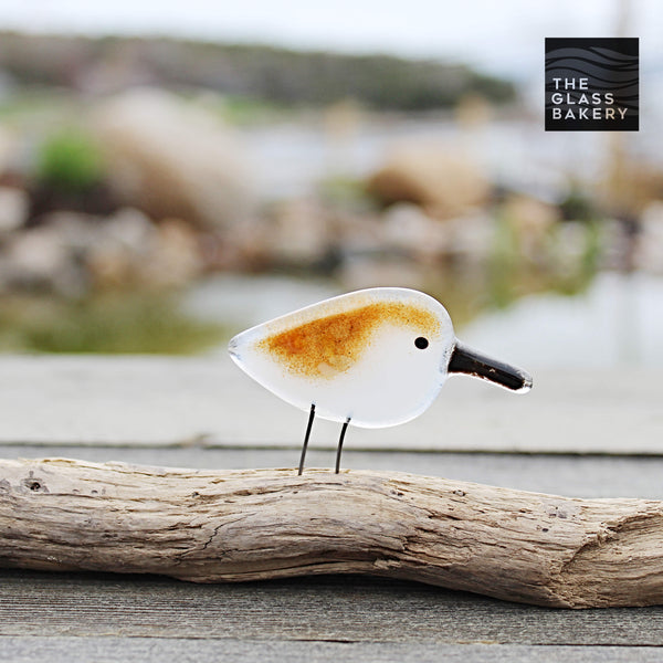 Load image into Gallery viewer, Baby brown and white sandpiper made from glass, sits on driftwood. In the background is a watery scene.
